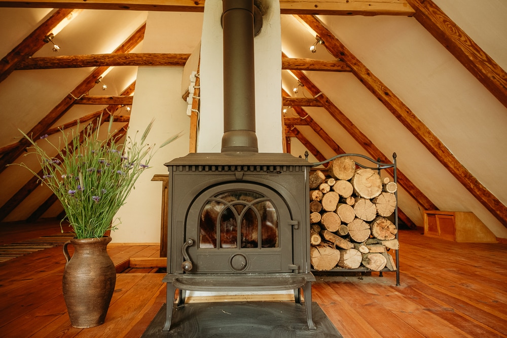 A rustic wood-burning stove is positioned against a wall of stacked firewood. A pottery vase, filled with dried flowers, complements the warm wooden beams and flooring of the spacious attic area, creating a cozy and inviting focal point.