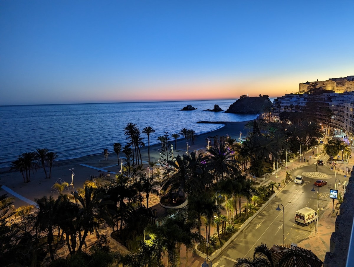 The image depicts a serene coastal view at twilight, with a smooth sea reflecting hues of orange and purple in the sky. Palm trees line the beach, while a quiet road is visible, leading to the gently lit buildings in the background.