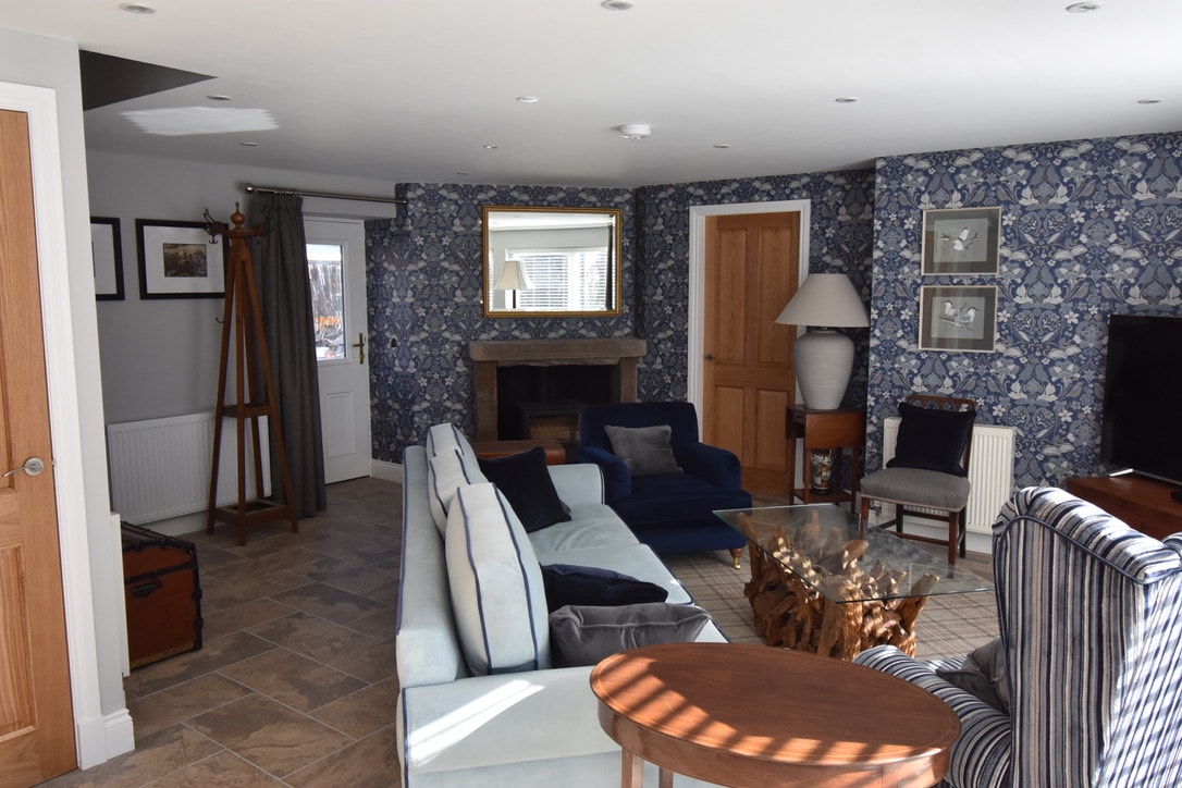 An open living area features a combination of seating, including a large sectional sofa and accent chairs. A coffee table made of natural wood rests on tiled flooring. Decorative wallpaper adds a patterned touch, while a fireplace and large windows fill the space with light.