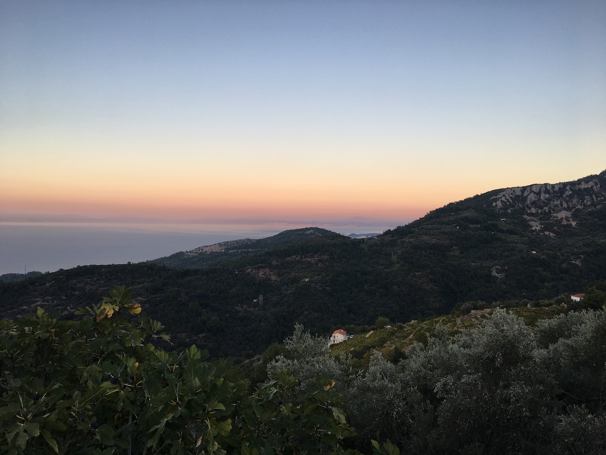 A panoramic view showcases rolling hills and lush greenery at dusk, with soft pastel hues of pink, orange, and blue lingering in the sky. The distant sea is visible on the horizon, blending subtly into the landscape.