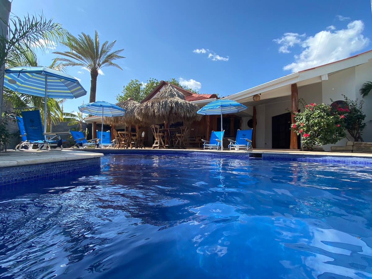 A large swimming pool reflects the clear blue sky, surrounded by palm trees and sun loungers. Shaded seating is provided under a thatched roof gazebo in the background, with colorful umbrellas offering additional shade. The inviting space is designed for relaxation.