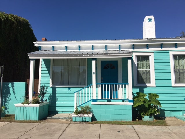 The exterior of a charming cottage features a vibrant light blue color. A welcoming porch with white railings and a bright blue door enhances the inviting appearance. Planters with plants and flowers are situated on either side of the entrance, contributing to the cheerful curb appeal.