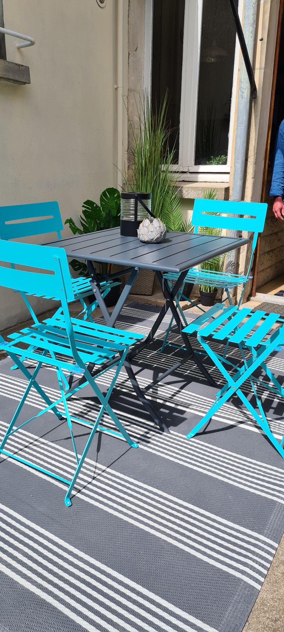 An outdoor seating area is presented with a small black table surrounded by four turquoise folding chairs. A lantern with a decorative item sits at the center of the table. A striped outdoor rug complements the scene, with greenery partially visible in the background.