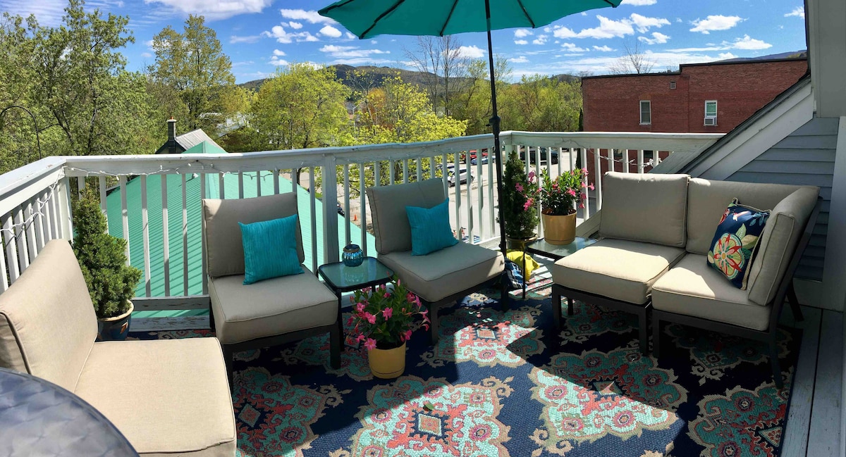 A private deck area is furnished with comfortable seating and colorful throw pillows. A small table sits in the center, surrounded by potted plants and vibrant flowers. An umbrella provides shade, enhancing the inviting outdoor space with a scenic view of the surrounding trees and buildings.