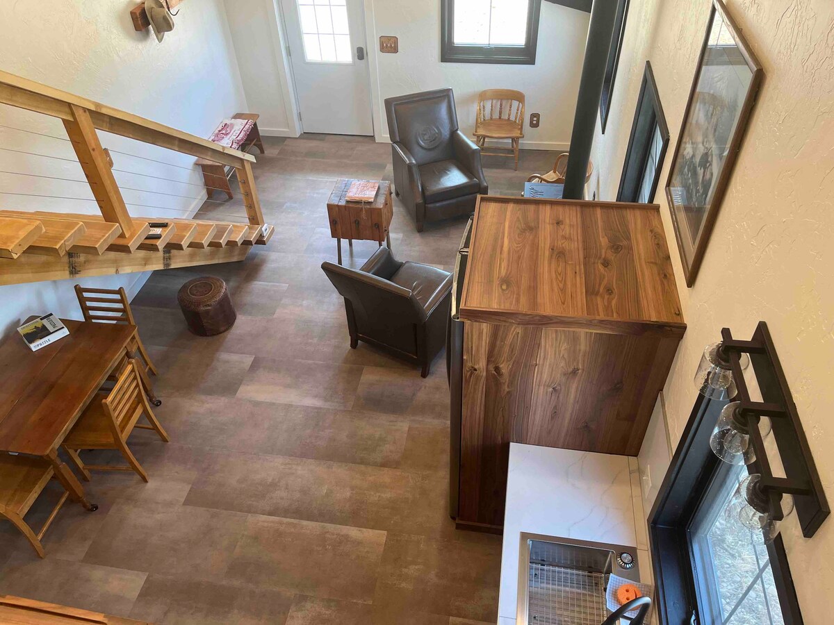 An open living space is highlighted by wooden accents and natural light. A dining table with four chairs sits alongside a comfortable leather armchair. A rustic cabinet is positioned near a small kitchenette featuring a sink. A staircase leads to the lofted area.
