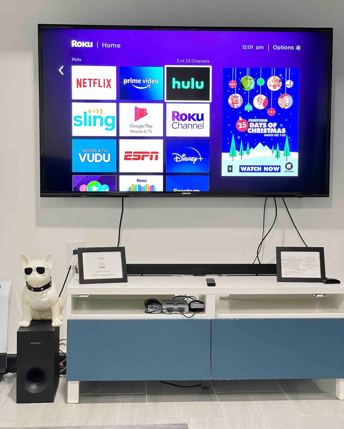 A large smart TV is mounted on the wall, displaying various streaming services such as Hulu and Netflix. Below, a modern media console in blue and white houses a sound system and decorative items. A whimsical dog figurine sits beside the television.