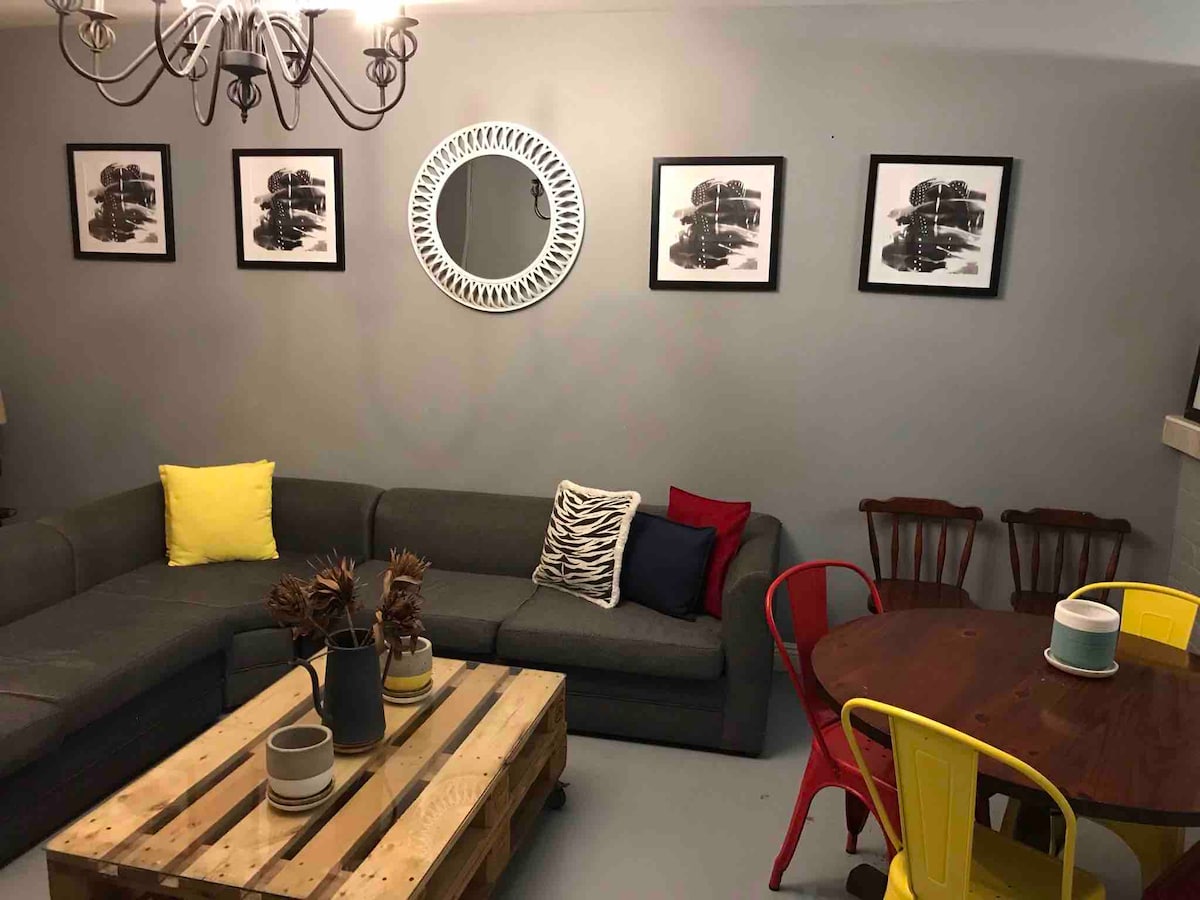A cozy living area features a large gray sectional sofa adorned with colorful cushions. A wooden pallet coffee table is surrounded by a round table and chairs in vibrant yellow and red. Black-and-white artwork decorates the walls, complemented by a decorative mirror.