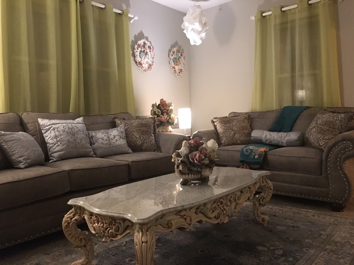 The living room features two comfortable sofas adorned with decorative pillows, complemented by a stylish marble coffee table. Soft lighting is provided by a lamp, and green curtains softly frame the windows, creating a calming ambiance. Decorative art pieces enhance the wall decor.