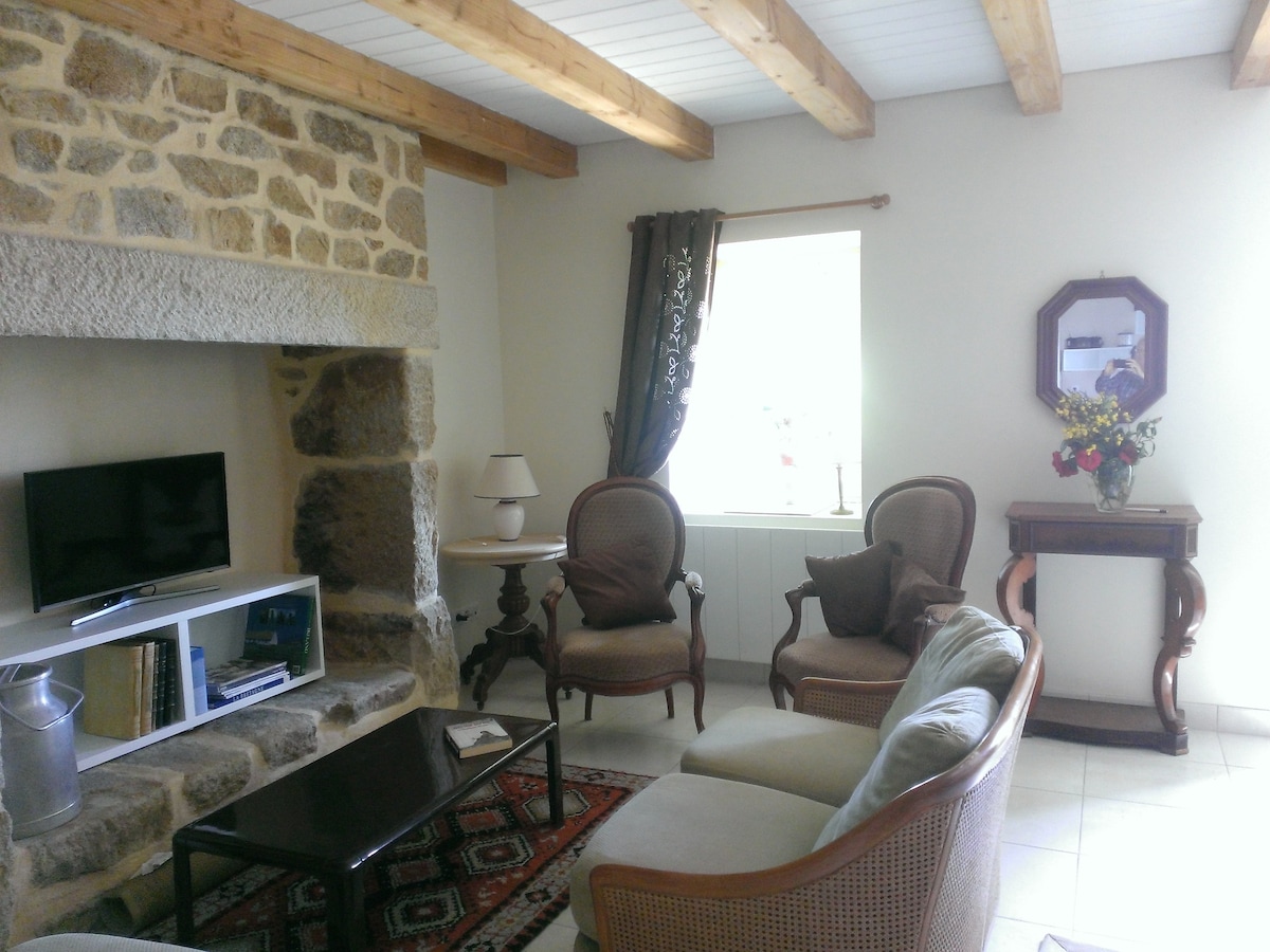 A cozy living area features a stone fireplace and wooden beams overhead. Seating is arranged with a light-colored sofa and vintage armchairs. A dark coffee table sits on a patterned rug. A window allows natural light, complemented by a decorative side table with a vase.