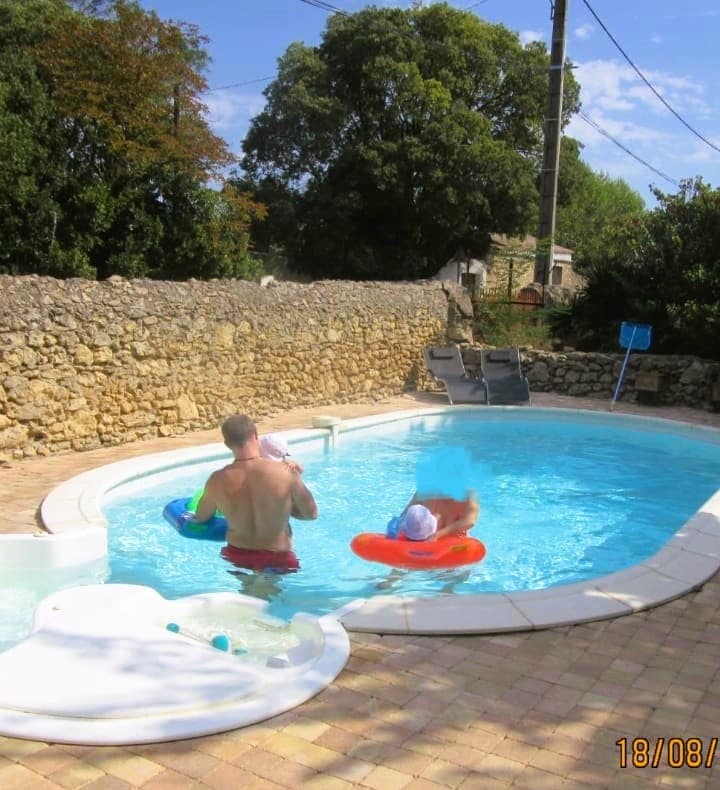 The outdoor swimming pool is surrounded by a natural stone wall and features a shallow area with a gentle slide. A parent is seen supervising two children floating in colorful inflatable tubes. Lush green trees provide shade nearby, creating a serene outdoor environment.