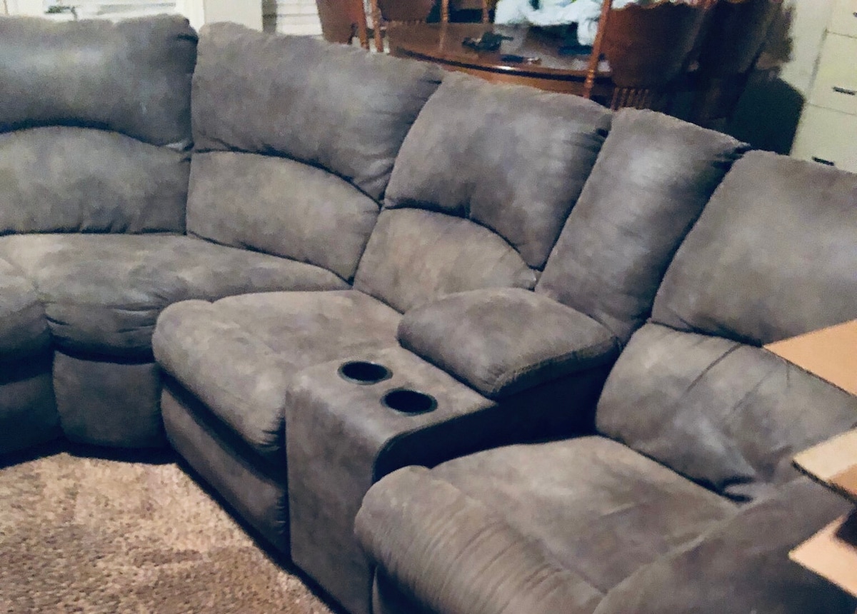 A comfortable sectional sofa in a neutral grey fabric is positioned in the living area. The sofa features a built-in armrest with cup holders, providing a space for relaxation. A portion of the dining area with wooden chairs and a table is visible in the background.