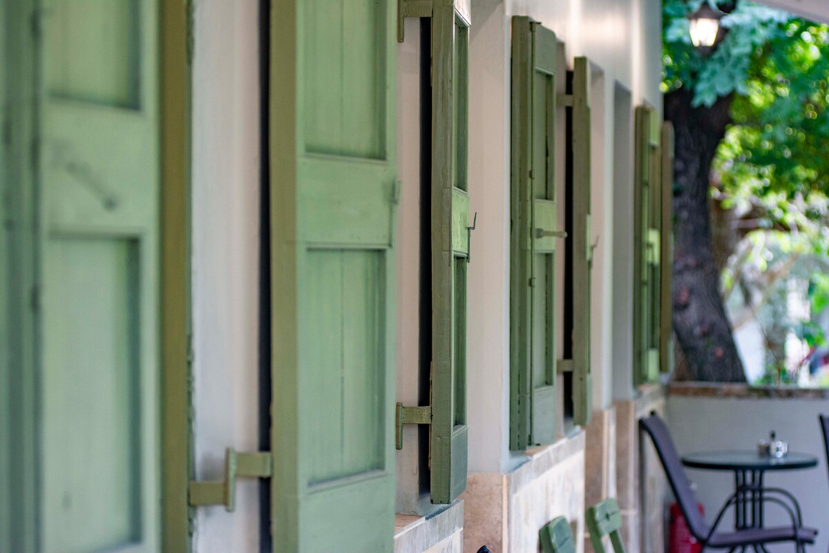 Lined green shutters are positioned along the exterior wall, framing the windows of the residence. A small outdoor seating arrangement is visible nearby, accompanied by natural greenery providing a serene atmosphere.
