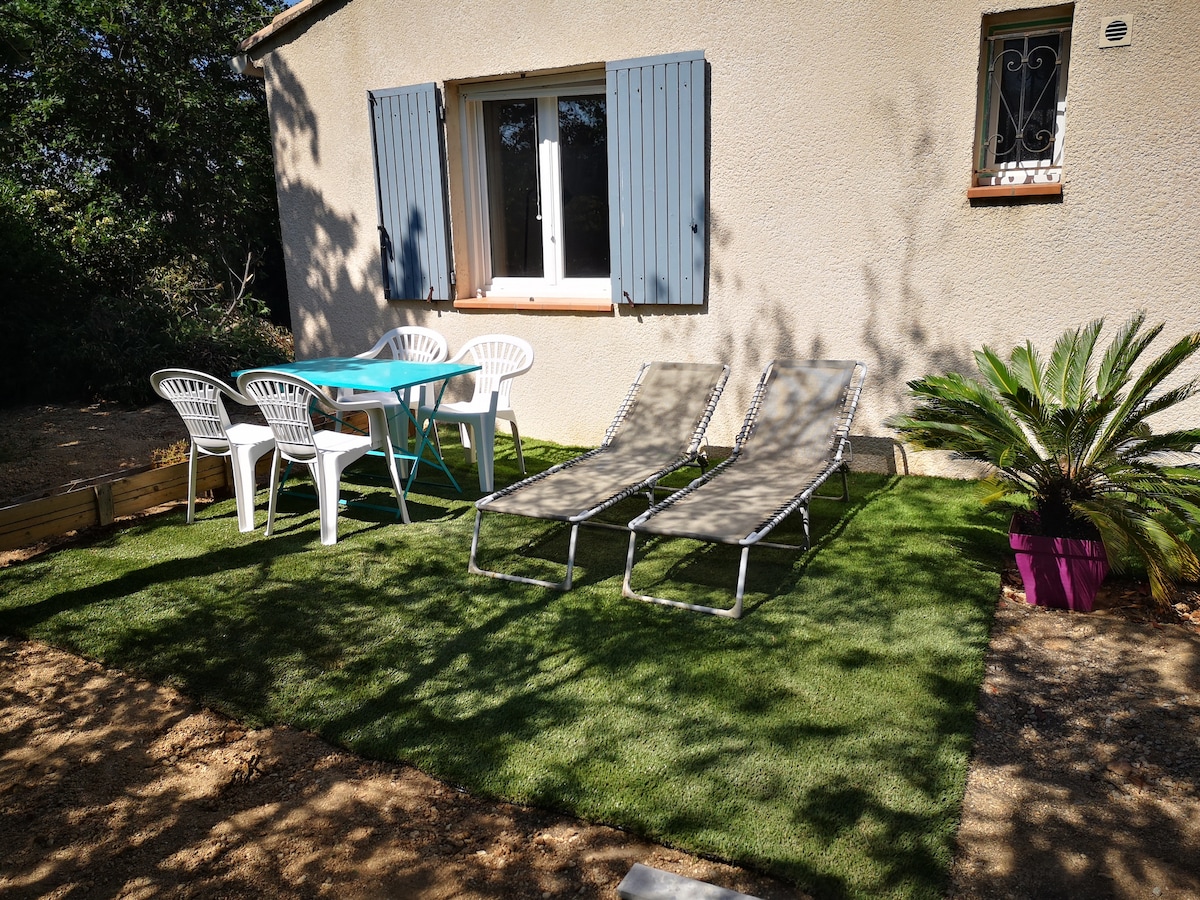 A private garden area features two lounge chairs and a small table surrounded by four white chairs. The well-maintained artificial grass enhances the outdoor space, while the house is adorned with blue shutters and a window allowing natural light to enter.