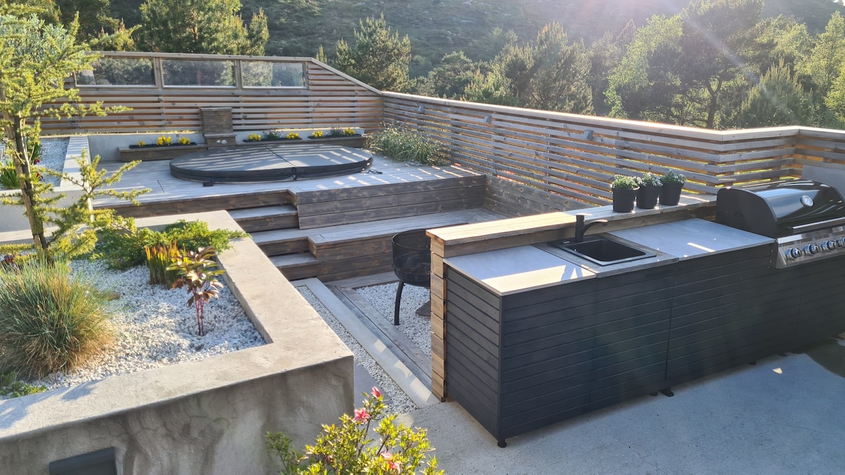 An outdoor area features a built-in outdoor kitchen with a grill and a seating area. Surrounding greenery and garden beds enhance the space. A hot tub is positioned on the wooden deck, offering a relaxing setting. Soft sunlight illuminates the area, creating a serene atmosphere.