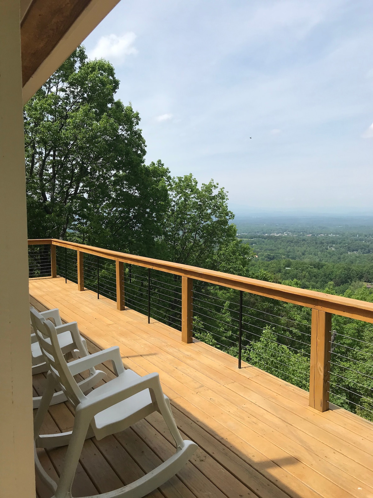 A wooden deck features multiple rocking chairs, inviting relaxation while enjoying expansive views of the surrounding landscape. Lush trees frame the horizon, and soft, natural light enhances the warm tones of the deck material.