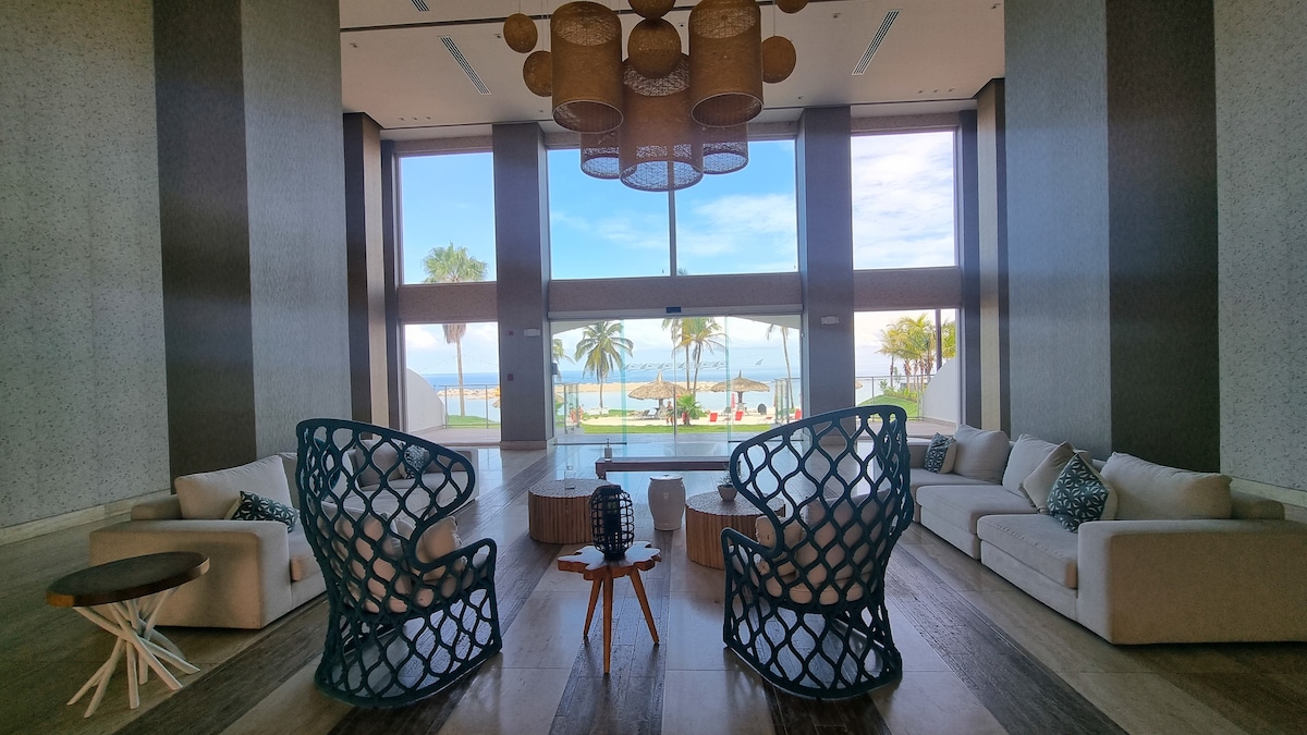 A spacious lounge area is designed with modern furniture, featuring comfortable sofas and unique woven armchairs. Large windows offer a clear view of the beach and palm trees. Natural light fills the space, creating an inviting atmosphere for relaxation.