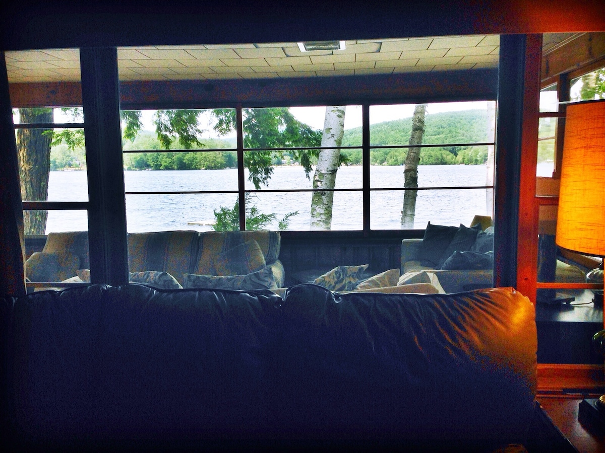 A cozy living area features large windows that frame views of the lake and surrounding trees. Soft seating, including sofas, is arranged for comfort, while a warm lamp provides gentle illumination. The peaceful lake scenery enhances the serene environment.