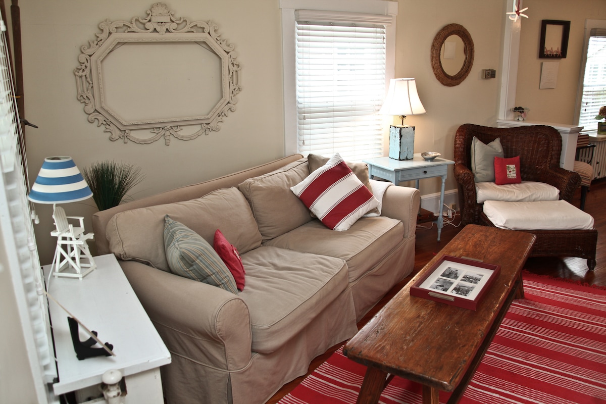 A comfortable living area features a beige sofa adorned with striped and patterned cushions. A rustic wooden coffee table sits in front, complemented by a cozy armchair. Natural light filters through white blinds, while a decorative framed piece hangs on the wall.