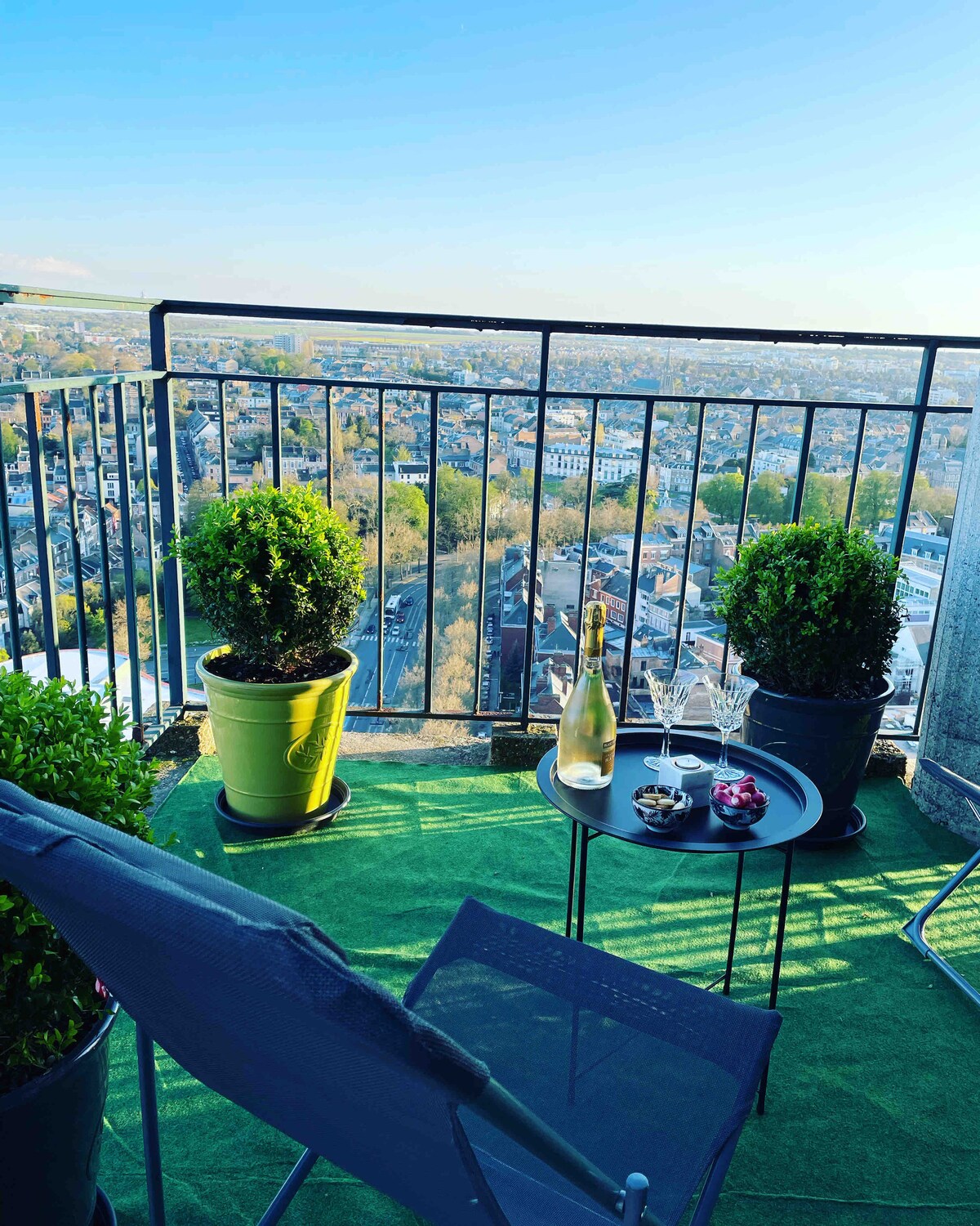 The terrace showcases a panoramic view of the city, framed by green planters. A small table holds a bottle of champagne and glasses, accompanied by a bowl of snacks. A comfortable chair invites relaxation, with vibrant greenery enhancing the outdoor space.