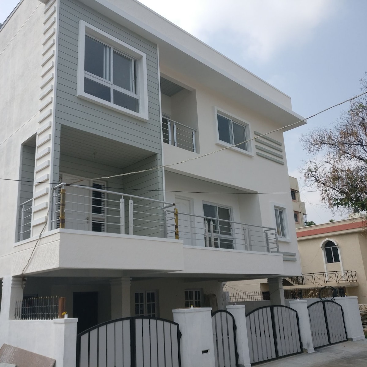 A modern three-story building features a light-colored facade with horizontal accents. Balconies with railings are visible on the upper levels, and large windows allow natural light to enter. A gated entrance leads to the parking area, providing secure access to the property.