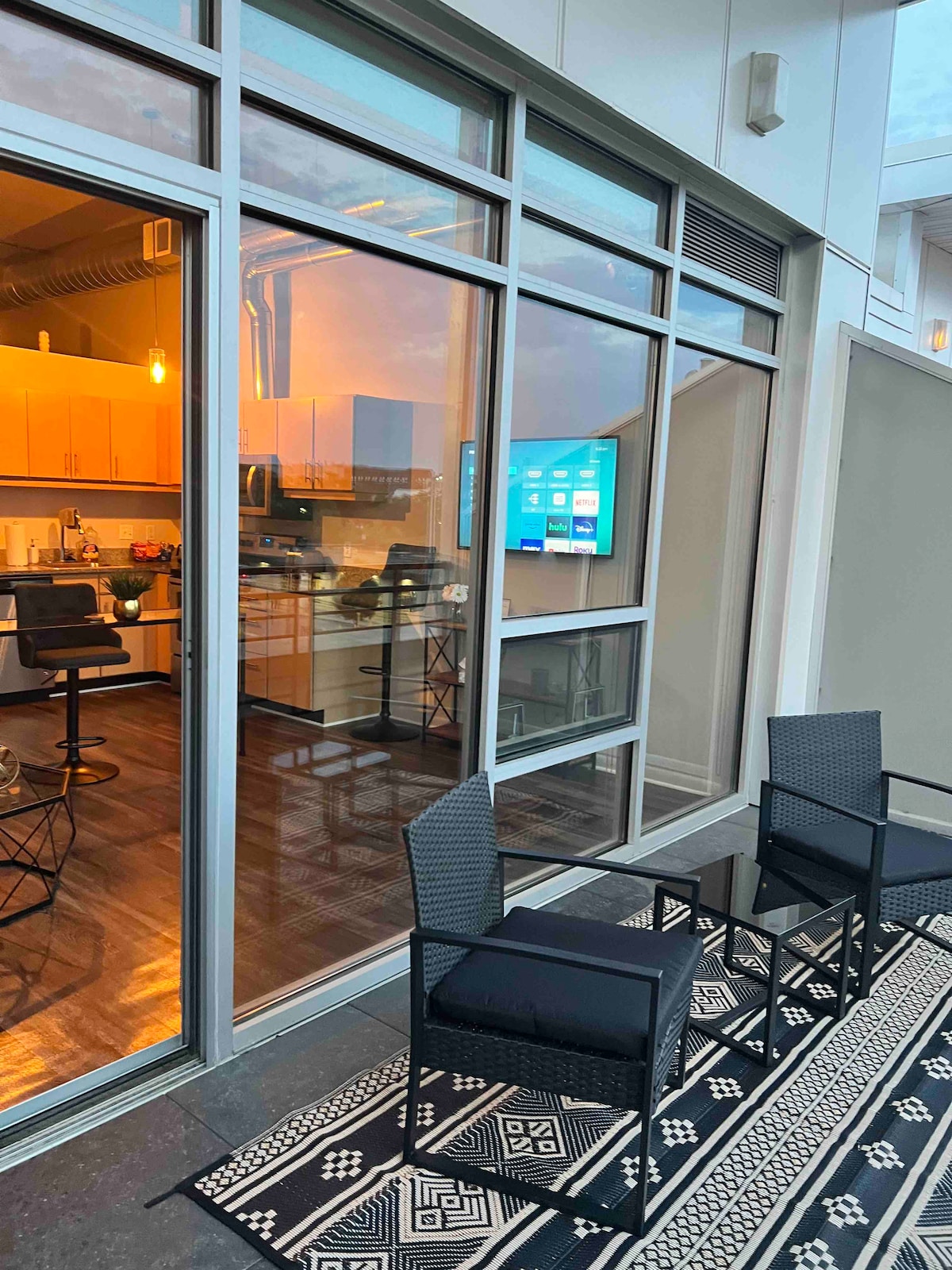 Two sleek black chairs are positioned on a patterned outdoor rug, set against large glass doors leading to a modern kitchen space. Inside, warm lights illuminate the kitchen area, complemented by a wall-mounted television. The space exudes a contemporary and inviting atmosphere.