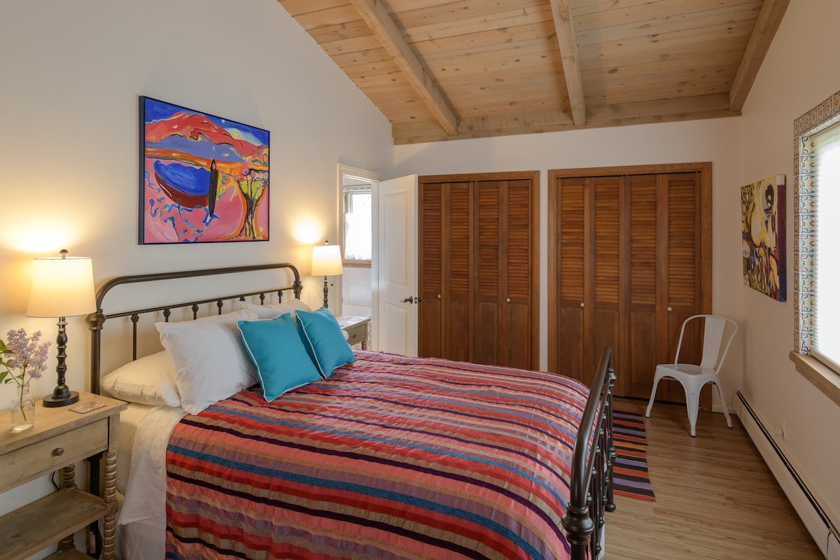 A spacious bedroom is furnished with a metal bed featuring a striped blanket and accent pillows. Natural light enters through a large window near a closet with wooden doors. Colorful artwork adorns the walls, and a small side table holds a vase with flowers.