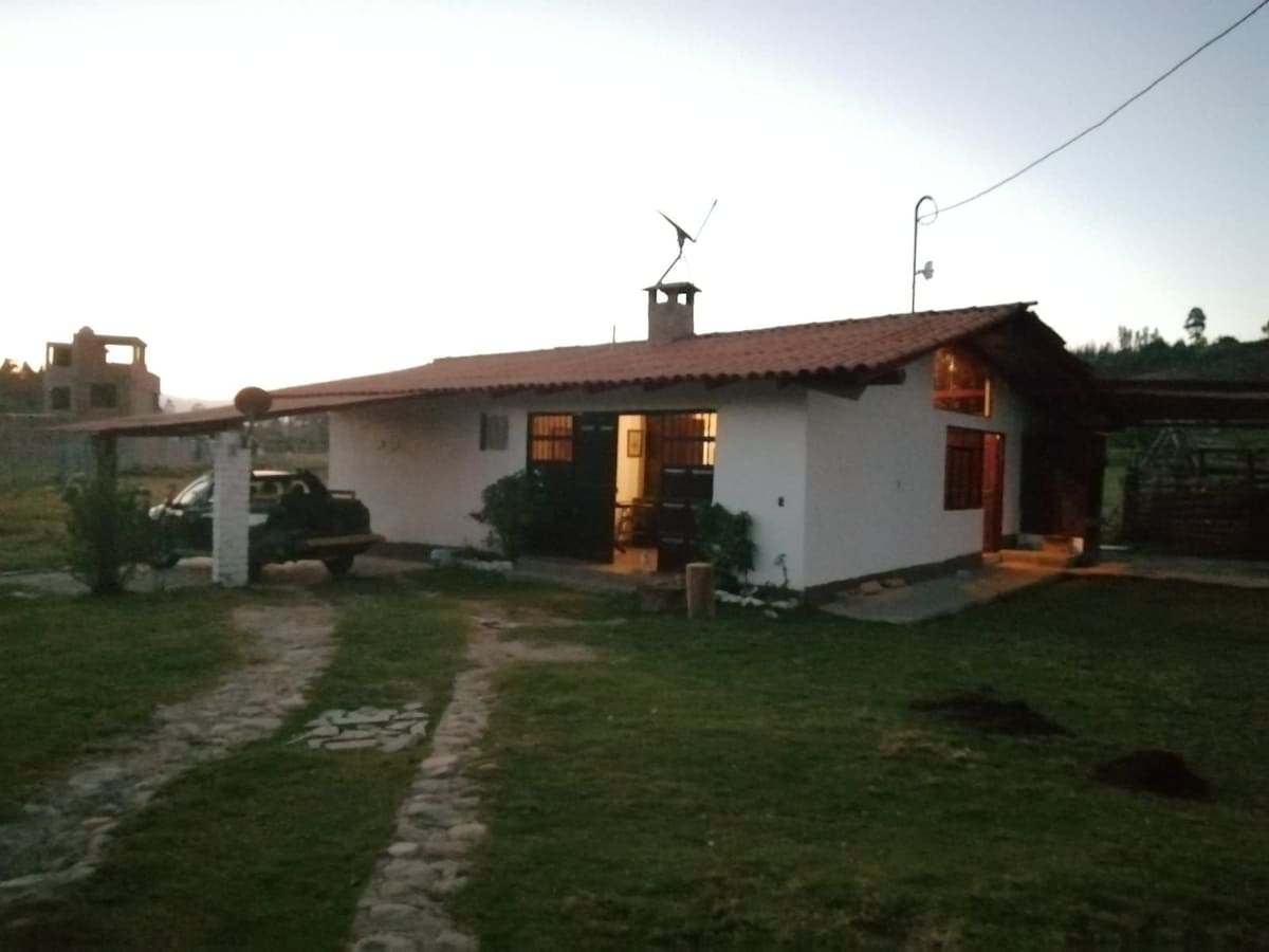 A spacious house is presented with a red-tiled roof and multiple windows, set against a serene twilight backdrop. A path of stones leads to the entrance, with a vehicle parked nearby. Lush greenery surrounds the home, complementing the peaceful country setting.