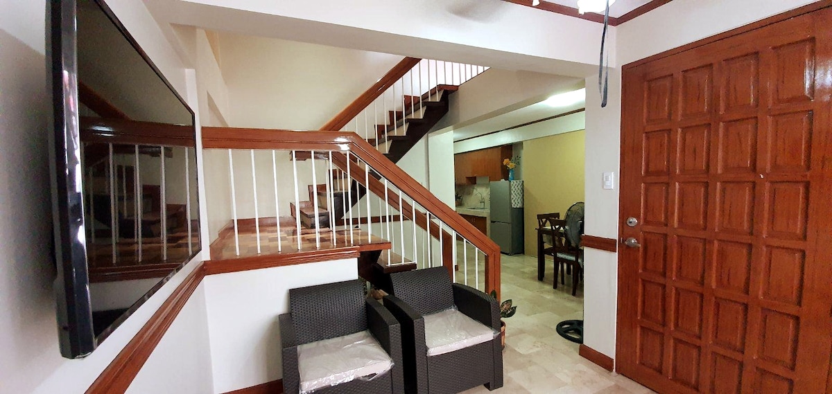 The entrance area features a welcoming layout with a stylish staircase leading to the upper floor. Two dark wicker chairs are positioned to the side, along with a mounted flat-screen television on the wall. Natural light fills the space, enhancing its inviting atmosphere.