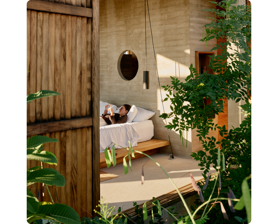 Two large, wood-slatted windows open to reveal a modern, concrete interior. A person lying on a platform bed with white linens watches their phone as daylight spills in from an unseen source. Greenery surrounds the space outside.