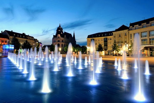 A vibrant water fountain display is captured at night, with streams of water illuminated in blue and white. The surrounding architecture features historical buildings under a sunset sky, creating a scenic urban environment.