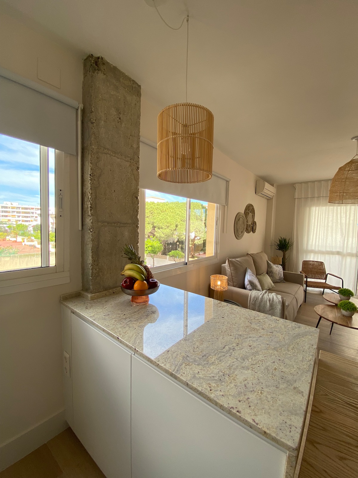 A bright kitchen area features a smooth granite countertop and a woven pendant light above. Natural light filters in through large windows, showcasing views of greenery outside. A cozy seating arrangement is visible, complemented by decorative elements and plants providing a relaxed ambiance.