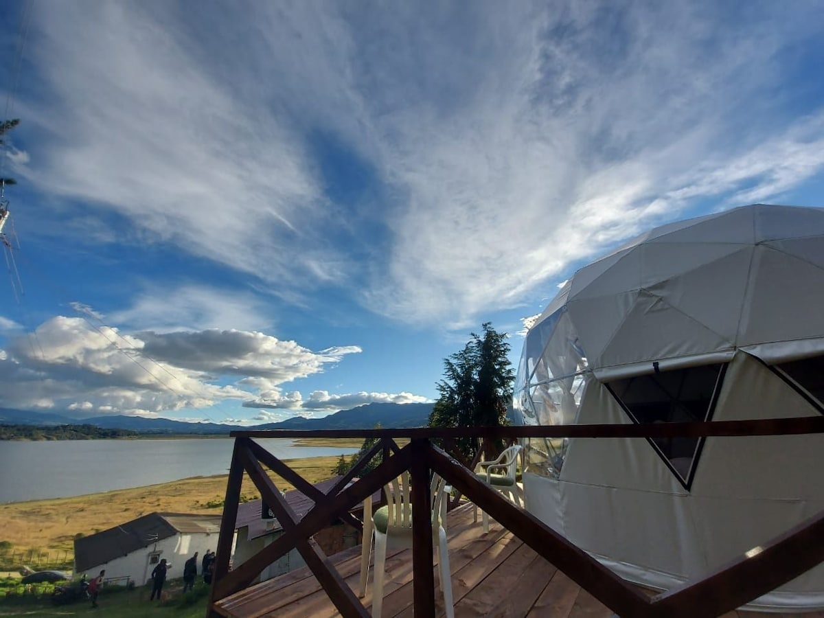 A geodesic dome structure is positioned on a wooden deck with a view of a serene reservoir. Expansive skies with scattered clouds are visible, while distant mountains create a tranquil backdrop. The surrounding landscape includes open fields and a few nearby buildings.