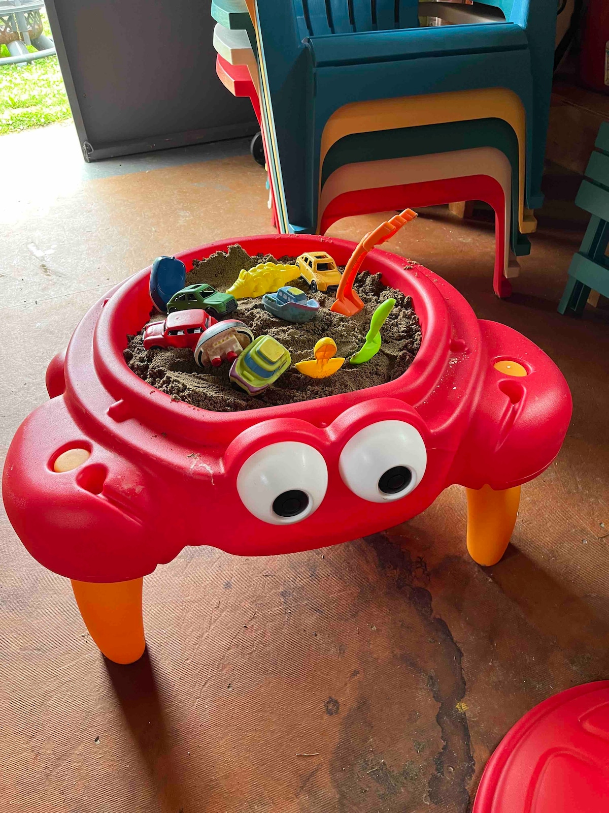 A brightly colored sand table shaped like a red crab is displayed, featuring various toys such as small vehicles and figurines. The table sits on four sturdy legs, with a playful design that adds a fun element to the space. Additional plastic chairs are visible in the background.