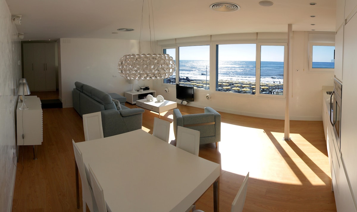 A bright living space features large windows offering views of the beach and ocean. A modern dining table is accompanied by white chairs. A comfortable seating area is arranged around a coffee table. Natural light floods the room, highlighting the airy, open layout.