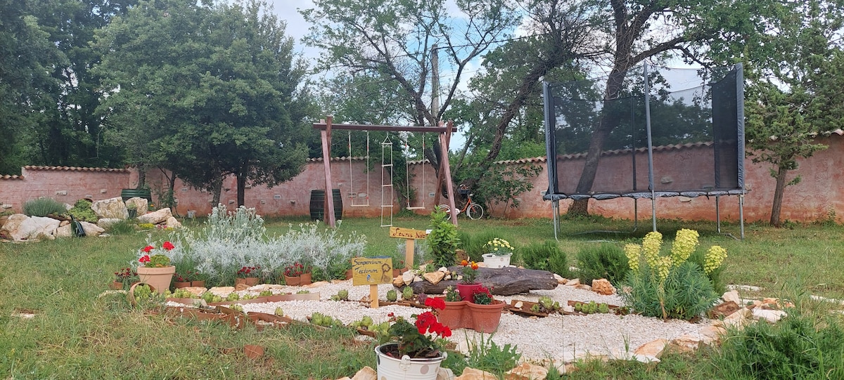 A spacious outdoor area is set within a fenced garden, featuring lush green grass and decorative flower beds. A small playground with swings and a trampoline is visible, surrounded by trees. Pathways made of stones and gravel connect various plants and decorative elements.