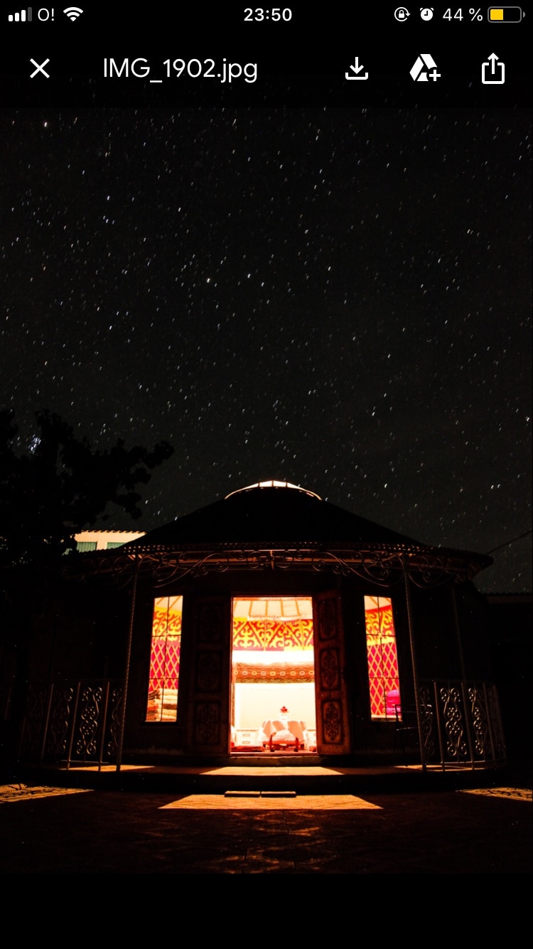 A traditional Kyrgyzstani yurt is illuminated from within, contrasting against a night sky filled with stars. The entrance showcases intricate patterns and warm light, welcoming visitors. The surrounding area is dark, emphasizing the yurt's vibrant and inviting atmosphere.