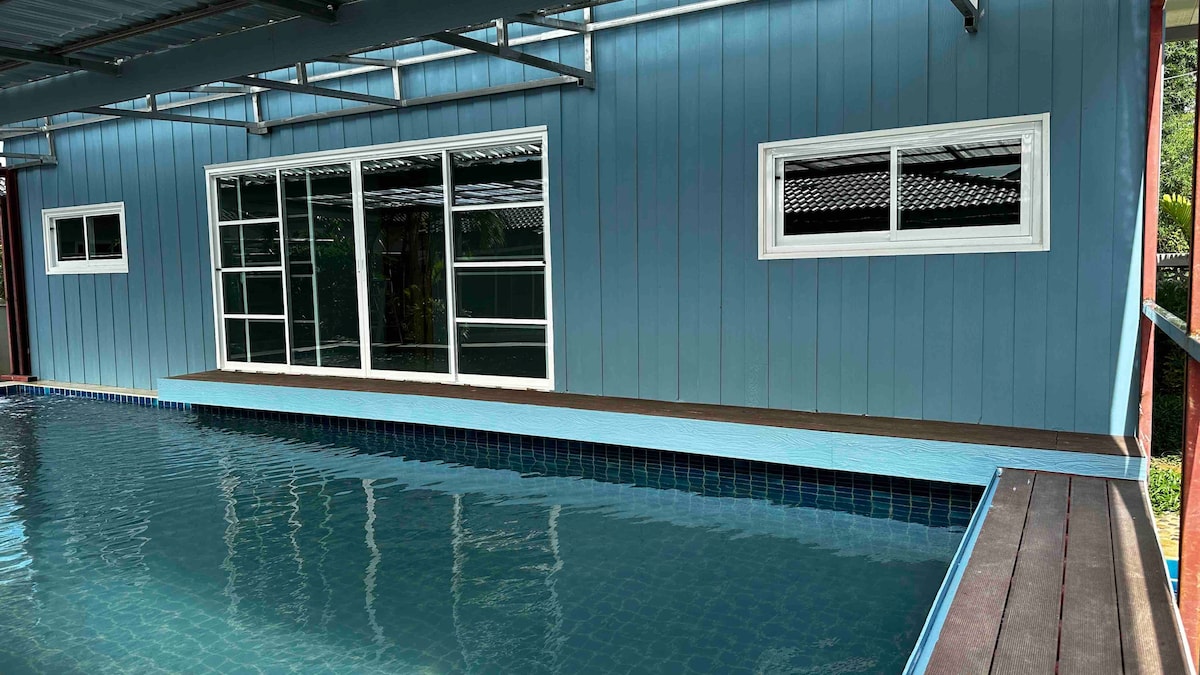 A serene pool area features a smooth surface of water reflecting light. The walls of the adjacent building are painted in soft blue, with large windows providing a view of the outdoor greenery. A wooden deck borders the pool, enhancing the outdoor experience.