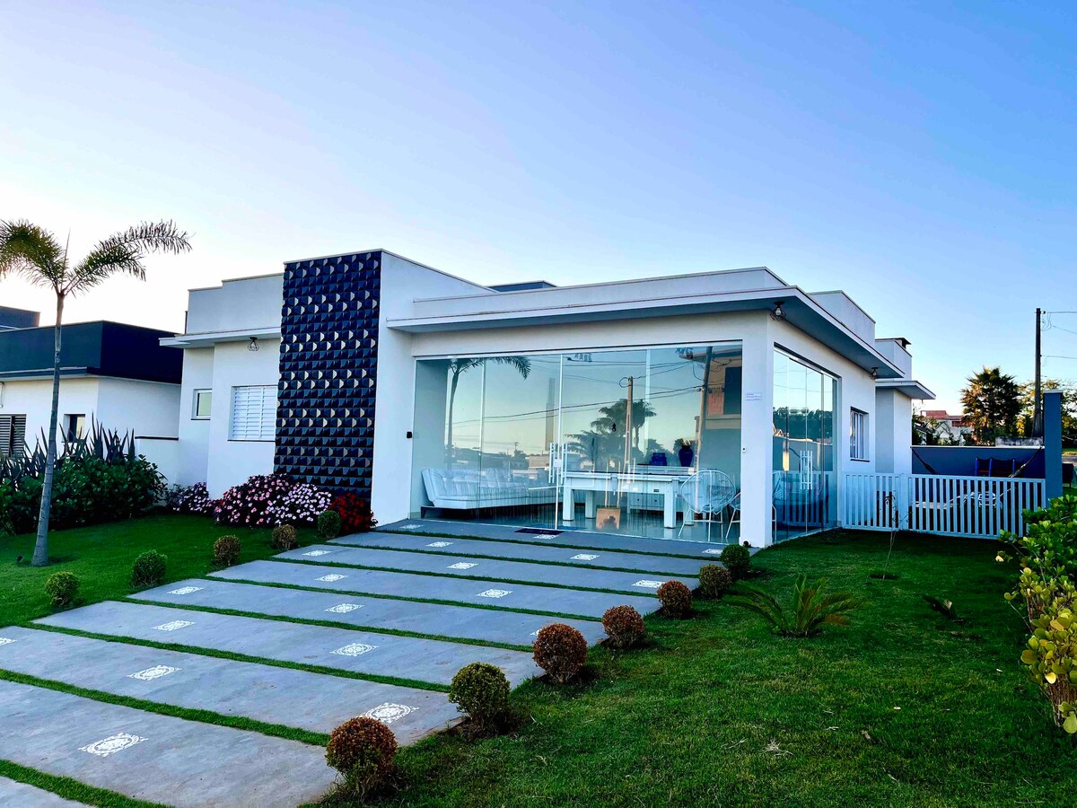 A modern single-story house is shown, featuring large glass windows that provide a view of the outdoor area. The lush green lawn is complemented by decorative hedges and shrubs. A paved pathway leads to the entrance, enhancing accessibility.