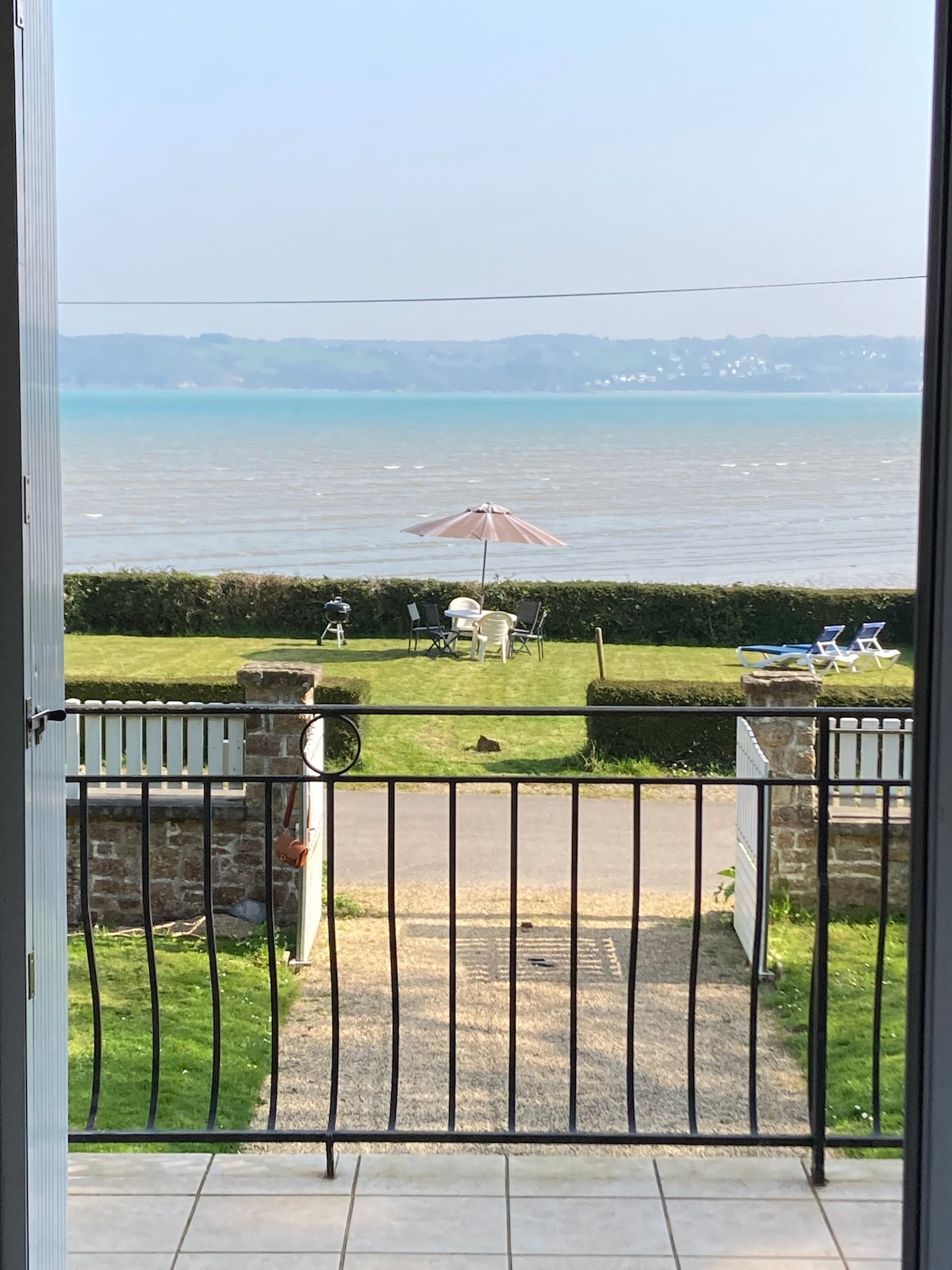 A view from the entrance features a well-maintained garden overlooking the water. White patio furniture is arranged under a parasol, with a manicured hedge and pathway leading to the shore, creating a serene outdoor space.