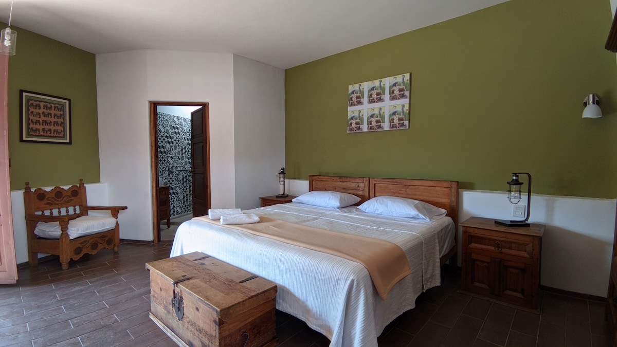A spacious room features a large bed with crisp linens, complemented by wooden bedside tables. A light-colored blanket drapes over the bed, and a cozy seating area with traditional wooden furniture is visible. Decor includes framed photos on a green wall.