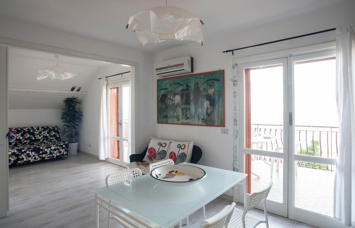 A bright living space features a dining table surrounded by white chairs, with natural light streaming through large glass doors. A colorful artwork adorns the wall, complemented by a patterned sofa in the adjacent area. Indoor plants add a touch of greenery.