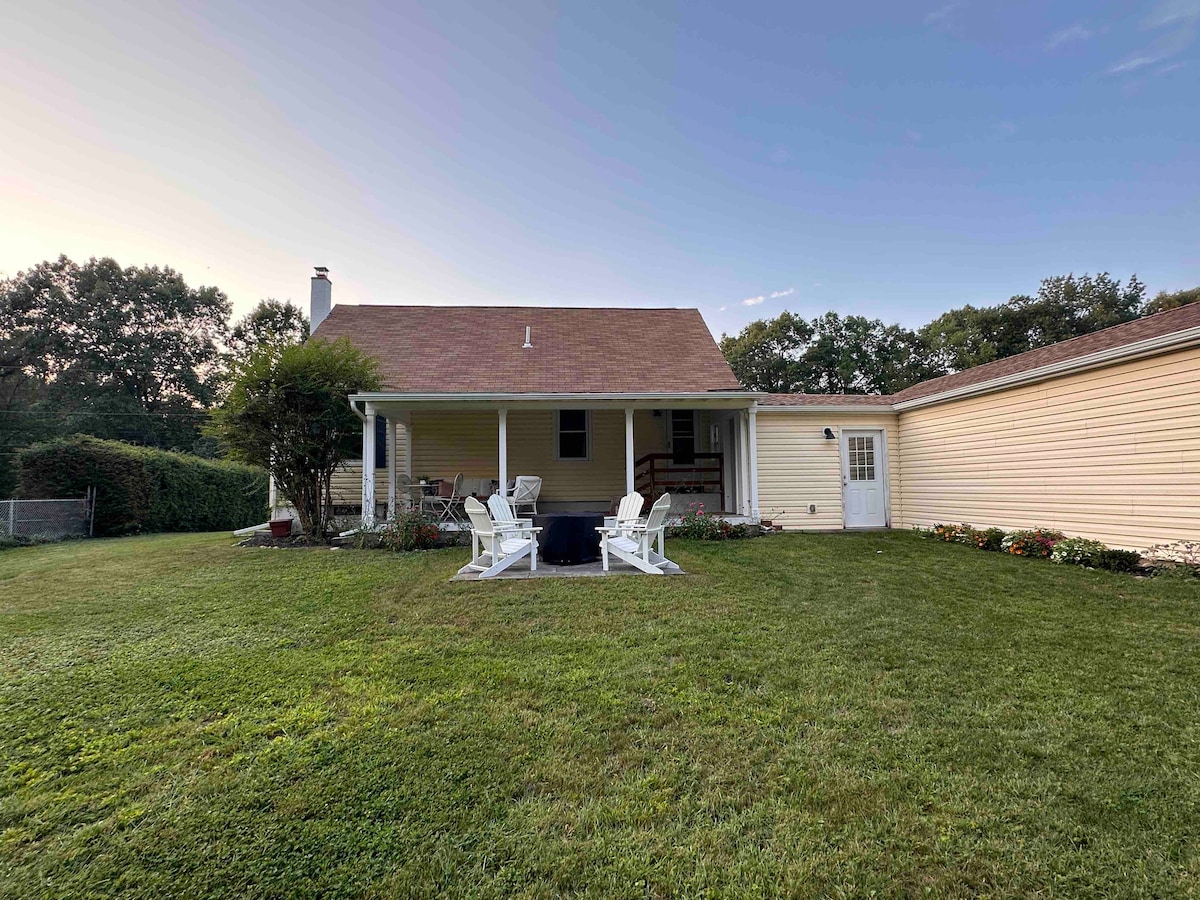 The backyard features a well-maintained lawn with several white Adirondack chairs arranged around a central fire pit. A covered porch provides shelter, while flowerbeds along the edges add color to the outdoor setting. The house has a warm yellow exterior, creating a welcoming atmosphere.
