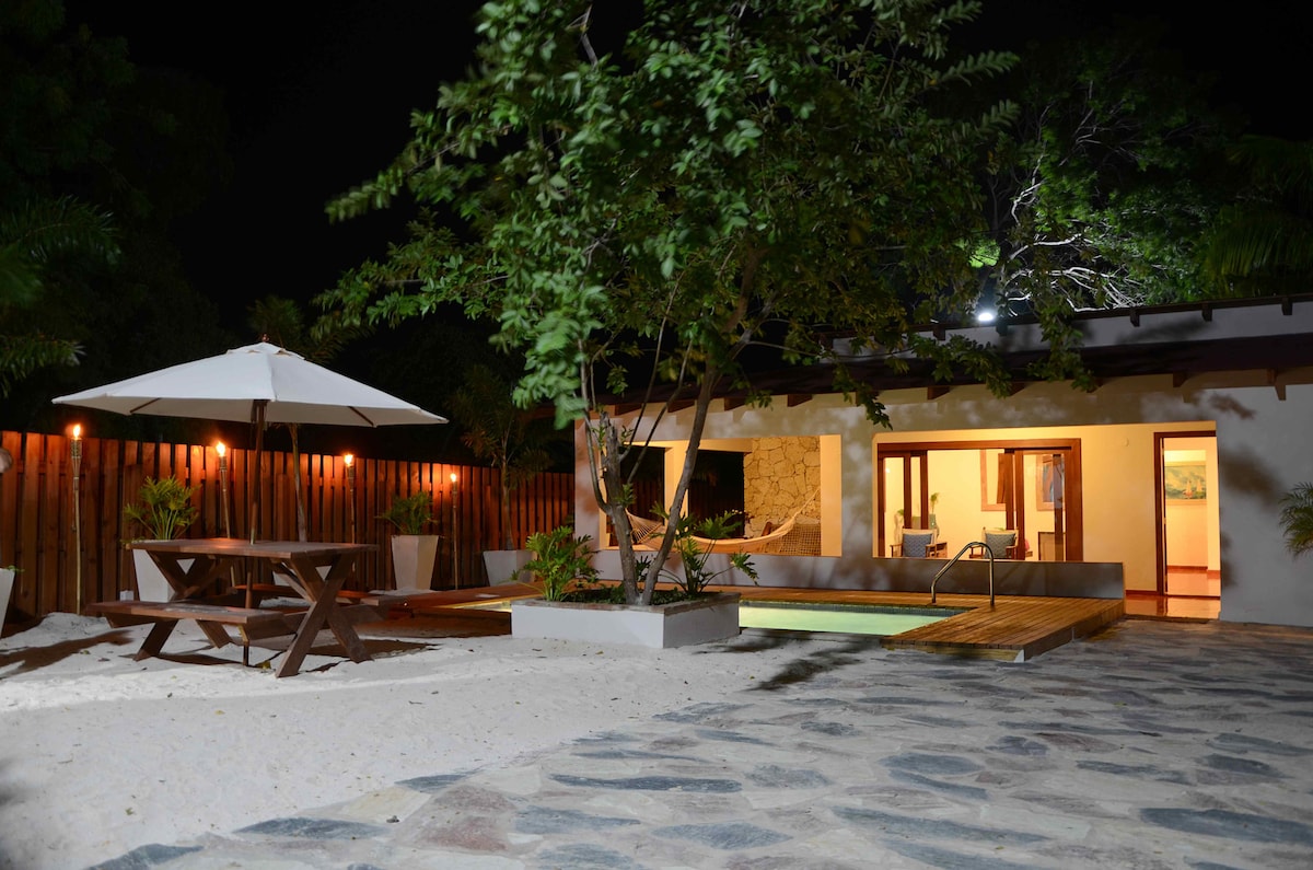 A private outdoor space is featured, showcasing a rustic wooden dining table beneath an umbrella, surrounded by soft sandy areas. A lighted pool and hammock are visible, with gently lit interiors peeking through glass doors, enhancing the tranquil ambiance of the villa at night.