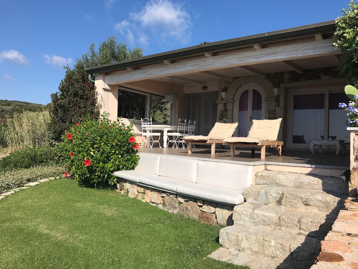 A charming outdoor terrace is presented, featuring a comfortable seating area with lounge chairs and a dining table surrounded by greenery. A well-maintained garden with blooming flowers and a manicured lawn provides a serene view, complemented by the natural landscape in the background.