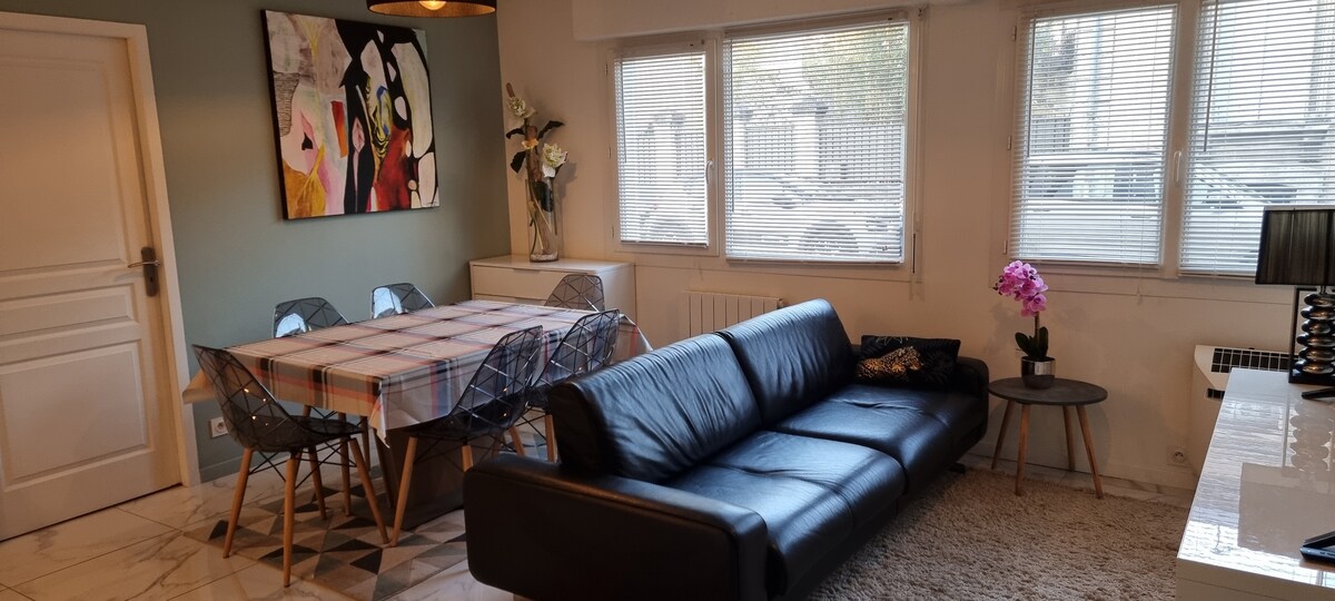 A bright living area features large windows allowing natural light to fill the space. A sleek black sofa is positioned opposite a dining table set for six. Colorful artwork adorns the wall, and a delicate vase with flowers adds a touch of elegance.
