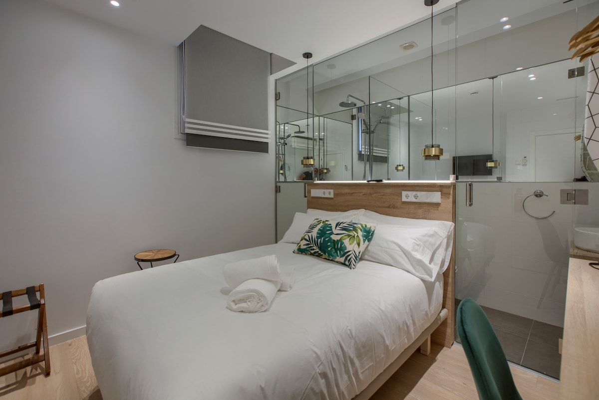 A modern room features a spacious bed adorned with decorative cushions. Mirrored walls enhance the sense of space, while soft lighting is provided by pendant lights. A stylish chair and a small side table add functional touches to the minimalist decor.
