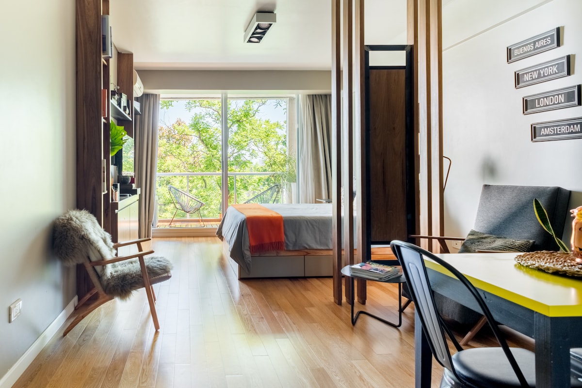 A well-designed living space features a spacious bed framed by large windows that allow natural light. A small dining table and chairs are present, alongside a seating area with a cozy armchair. Decorative accents enhance the modern aesthetic.