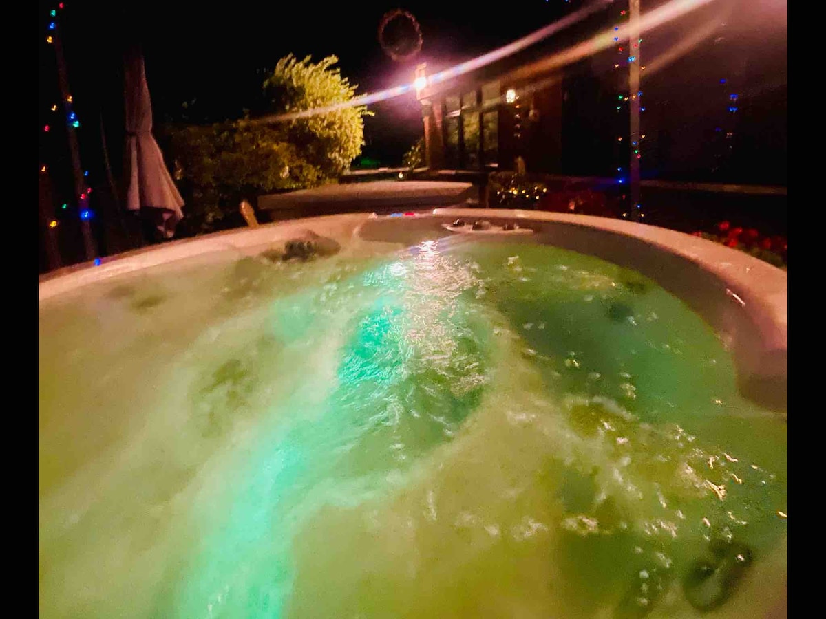 The hot tub is surrounded by intimate lighting and greenery, creating a tranquil atmosphere. The water appears inviting, with gentle bubbles and a soft glow from colored lights reflecting off the surface, enhancing the setting for a relaxing experience.