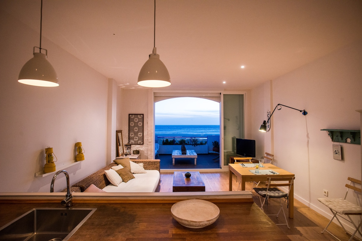 A spacious living area is presented with a comfortable sofa and a wooden coffee table, offering a view of the ocean through large sliding glass doors. A dining table and chairs are visible, along with modern lighting fixtures, creating an inviting space for relaxation.