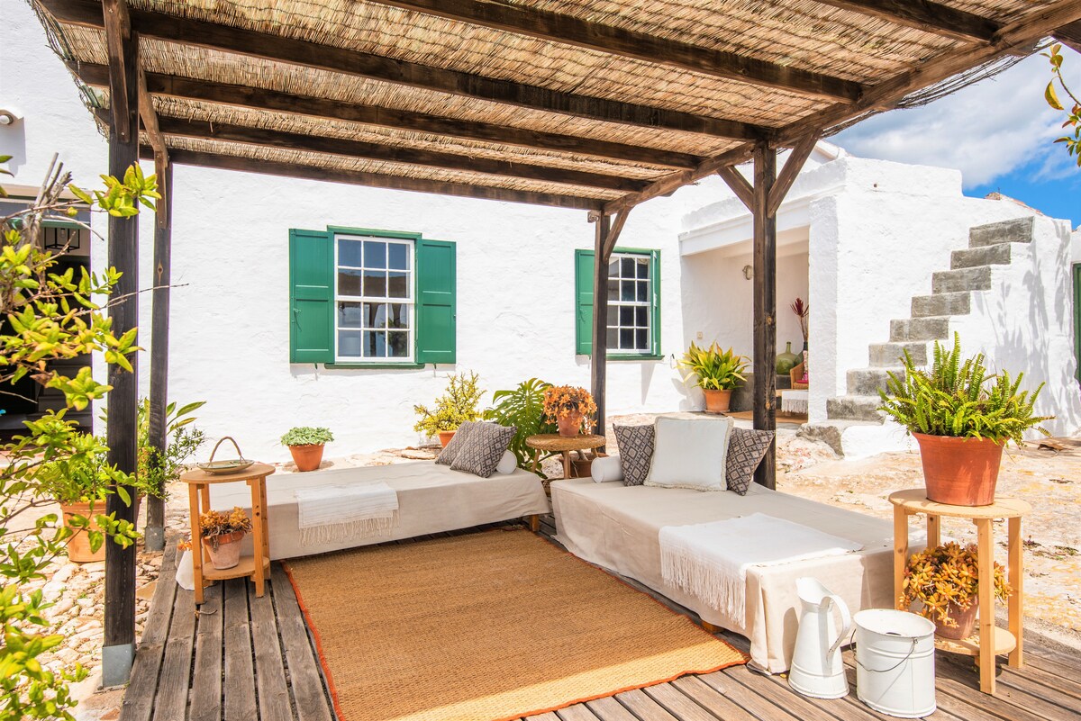 A shaded outdoor area features two lounging spaces adorned with soft cushions. A natural fiber rug lies beneath, surrounded by potted plants and a wooden lantern. White walls and green shuttered windows create a charming backdrop, enhancing the inviting setting.
